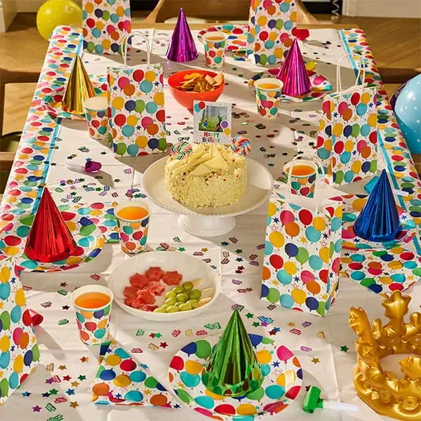 Outdoor birthday party table with a birthday cake, colourful tableware, party hats, gift boxes, balloons, and birthday decorations.